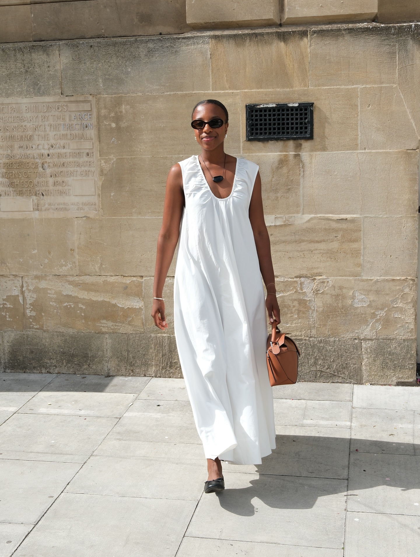 British Taffy Msipa poses in black oval sunglasses, a black cord pendant necklace, ruched white sleeveless dress, tan Loewe Puzzle bag, and black ballet flats.