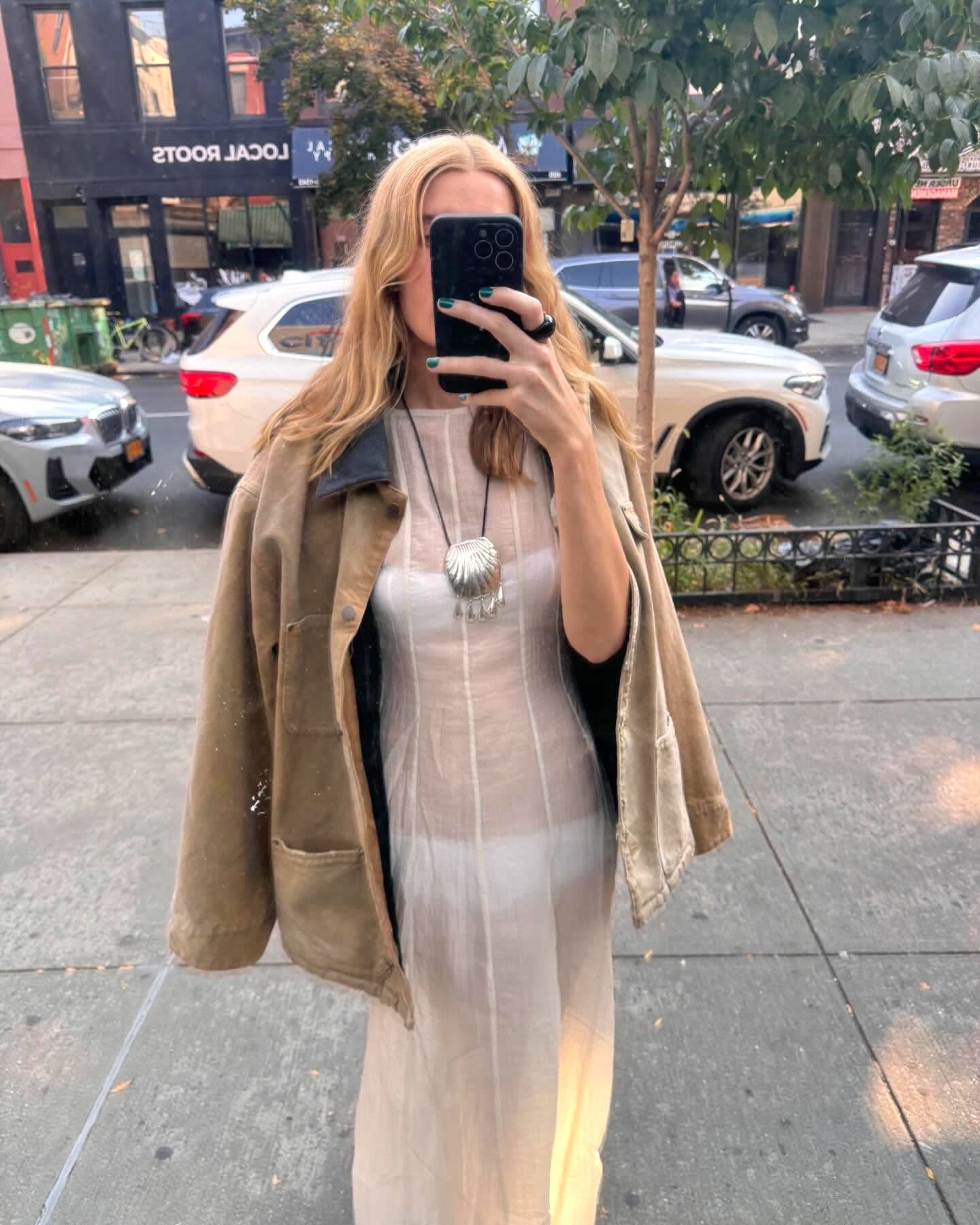 NYC-based style influencer Courtney Grow poses for a window selfie wearing a canvas barn jacket, a sheer white dress, and a silver Juju Vera Petra shell necklace.