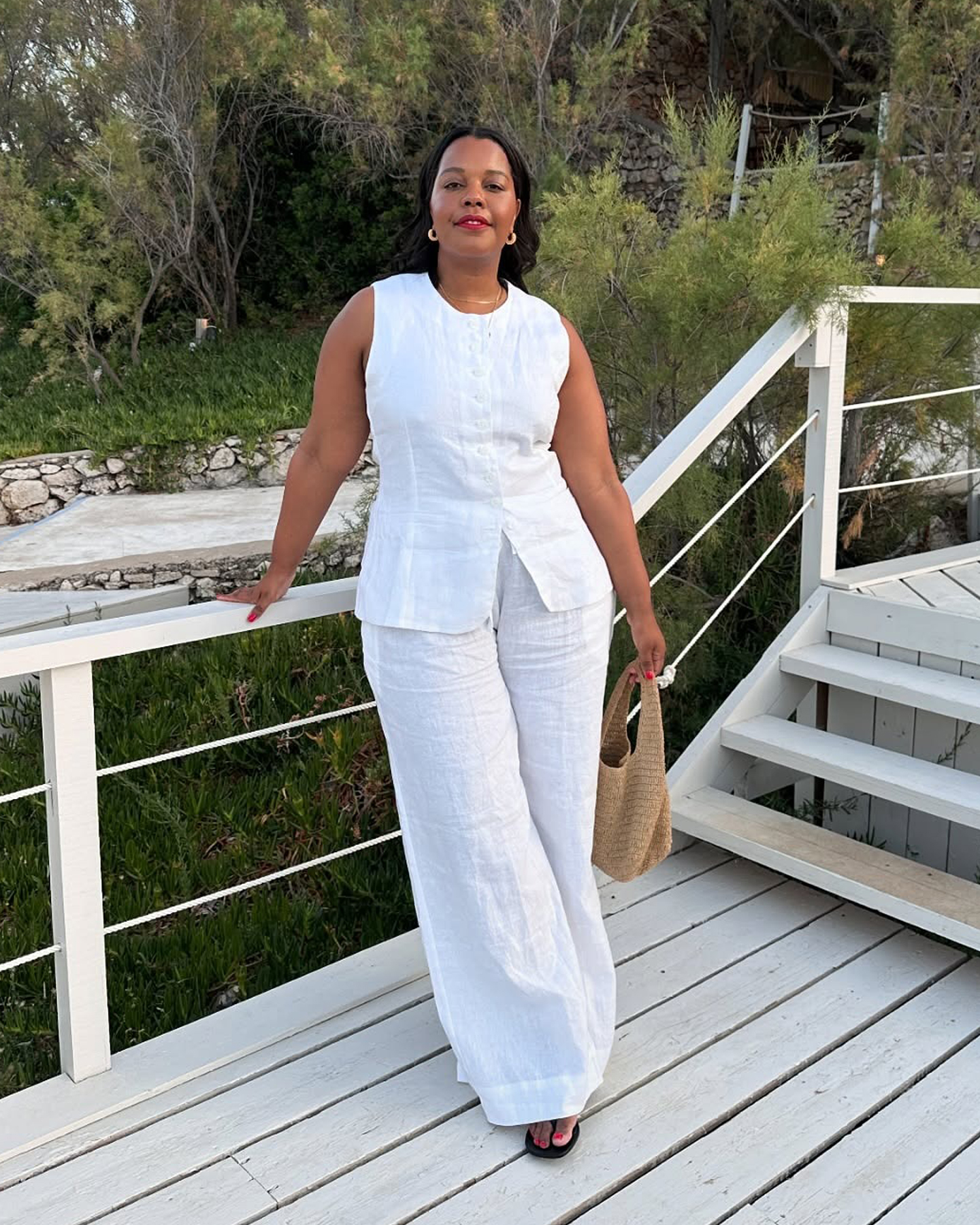 British influencer Karina Marriott poses at the The Peligoni Club in Greece wearing a matching white linen vest and pants with a mini woven bag and black flip-flop sandals.